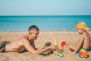 The family is playing on the beach. Son and father are playing with sand on the seashore. Dad with a child on the beach playing with toys. Summer time. Travel. photo