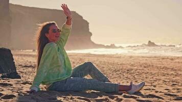 een meisje golven haar hand- zittend Aan de zand, een wild strand video