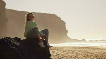 de flicka sitter på en stor sten, hår utvecklande i de vind, solnedgång på de hav video