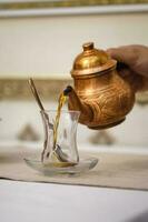 A man's hand pours tea into a Turkish cup from a copper teapot. photo