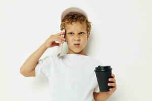 linda pequeño chico en un blanco camiseta gorra con un teléfono en un vaso con un bebida ligero antecedentes inalterado foto