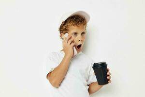 linda pequeño chico en un blanco camiseta gorra con un teléfono en un vaso con un bebida ligero antecedentes inalterado foto