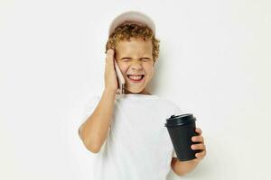 pequeño chico en un blanco camiseta gorra con un teléfono en un vaso con un bebida ligero antecedentes inalterado foto