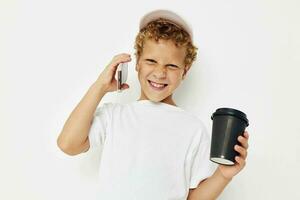 Photo portrait curly little boy in a white t-shirt cap with a phone in a glass with a drink light background unaltered