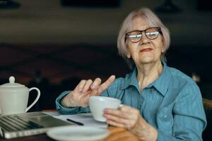 Portrait of an elderly woman with glasses sits at a table in front of a laptop Freelancer works unaltered photo