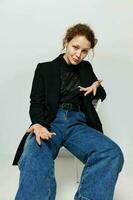 alegre mujer sentado en un silla en un negro chaqueta Moda posando ligero antecedentes inalterado foto