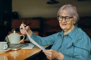 retrato de un mayor mujer con lentes se sienta a un mesa en frente de un ordenador portátil estilo de vida inalterado foto