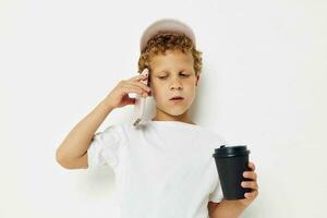 linda pequeño chico en un blanco camiseta gorra con un teléfono en un vaso con un bebida ligero antecedentes inalterado foto