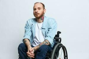 A man in a wheelchair looks thoughtfully at the camera, copy space, with tattoos on his arms sits on a gray studio background, the concept of health a person with disabilities, a real person photo