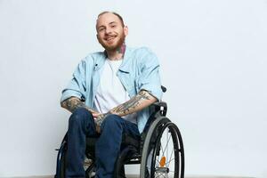 A man in a wheelchair and pointing a finger at, copy space, with tattoos on his hands sits on a gray studio background, health concept a person with disabilities, a real person photo