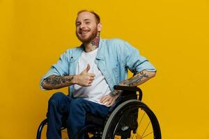 un hombre en un silla de ruedas sonrisa y felicidad, pulgar arriba, con tatuajes en su manos se sienta en un amarillo estudio fondo, el concepto de salud un persona con discapacidades foto