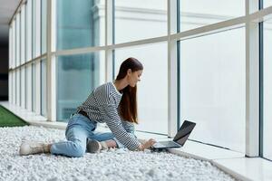 A woman sits in an office lobby by large windows and works on her laptop online via the Internet, freelance work anywhere in the world photo