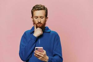 Portrait of a man with a phone in his hands doing looking at it and talking on the phone, on a pink background. Communicating online social media, lifestyle photo