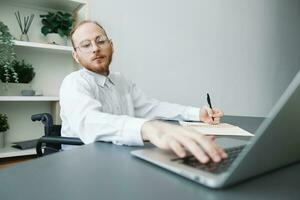 A man wheelchair businessman in the office works at a laptop, writing down ideas and a plan in a notebook, thoughtfulness, integration into society, the concept of working a person with disabilities photo