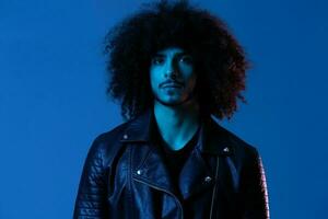 Portrait of fashion man with curly hair on blue background multinational, colored light, black leather jacket trend, modern concept. photo