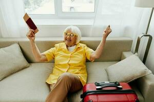 Happy senior woman with passport and travel ticket packed a red suitcase, vacation and health care. Smiling old woman joyfully sitting on the sofa before the trip raised her hands up in joy. photo