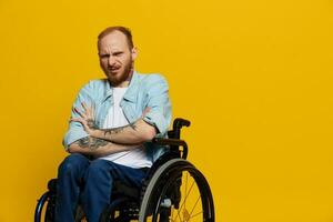 A man in a wheelchair problems with the musculoskeletal system looks at the camera is not satisfied, with tattoos on his arms sits on a yellow studio background, health concept man with disabilities photo