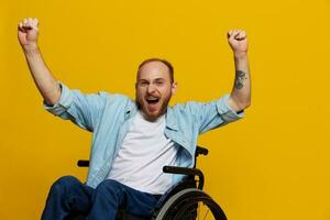 un hombre en un silla de ruedas sonrisa y felicidad, pulgar arriba, con tatuajes en su manos se sienta en un amarillo estudio fondo, el concepto de salud un persona con discapacidades foto