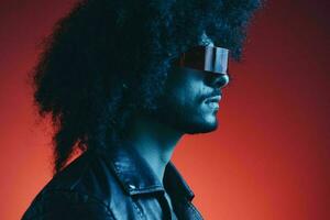 Portrait of fashion man with curly hair on red background with stylish glasses, multicultural, colored light, black leather jacket trend, modern concept. photo