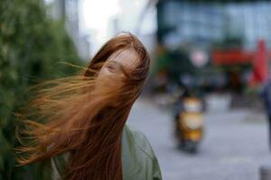 retrato de un hermosa joven mujer en el ciudad mirando dentro el cámara sonrisa con dientes con rojo volador pelo en de moda ropa en el ciudad en contra de bambú en primavera, estilo de vida en el ciudad foto