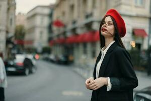Moda mujer retrato en pie en el calle en frente de el ciudad en elegante ropa con rojo labios y rojo boina, viajar, cinematográfico color, retro Clásico estilo, urbano Moda estilo de vida. foto