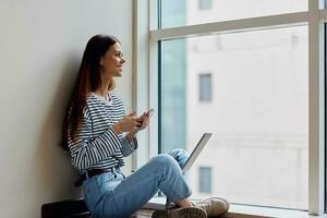 Happy woman sitting on the floor by the windows with a laptop on her lap and holding her phone, a view of the big city, the concept of work and learning online photo