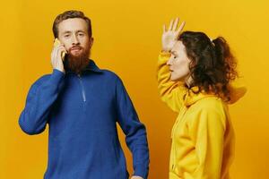 Man and woman couple with phone in hand call talking on the phone, on a yellow background, symbols signs and hand gestures, family quarrel jealousy and scandal. photo