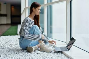 A woman smiling with teeth sits with a laptop by the large windows of a building in the city talking via video link through a computer, the concept of work and leisure in the big city photo
