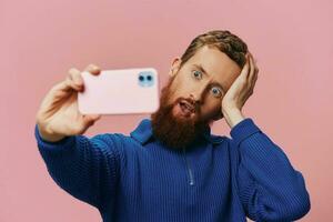 Portrait of a redheaded man with phone in hand taking selfies and photos on his phone with a smile on a pink background, blogger