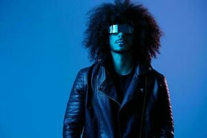 Portrait of fashion man with curly hair with stylish glasses on blue background multinational, colored light, black leather jacket trend, modern concept. photo