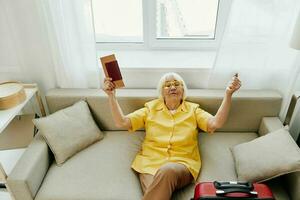 Happy senior woman with passport and travel ticket packed a red suitcase, vacation and health care. Smiling old woman joyfully sitting on the sofa before the trip raised her hands up in joy. photo