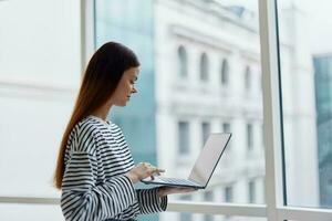 Woman with a laptop standing at the window in the office and looking out the window, work in an informal setting, business woman photo