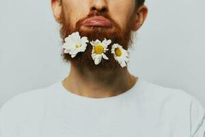 Portrait of a funny man in a white T-shirt with flowers daisies in his beard on a white isolated background, copy place. Holiday concept and congratulations. photo