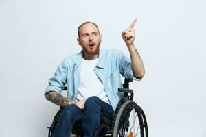 A man in a wheelchair and pointing a finger at, copy space, with tattoos on his hands sits on a gray studio background, health concept a person with disabilities, a real person photo