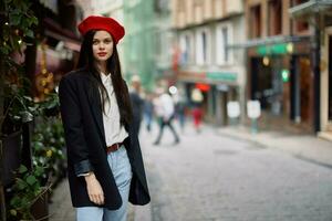 mujer Moda modelo soportes en el calle en el ciudad entre el multitud en un chaqueta y rojo boina y vaqueros, cinematográfico francés Moda estilo ropa, viaje a Estanbul foto