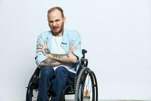 un hombre en un silla de ruedas mira a el cámara ira y agresión, con tatuajes en su brazos se sienta en un gris estudio fondo, salud concepto hombre con discapacidades foto