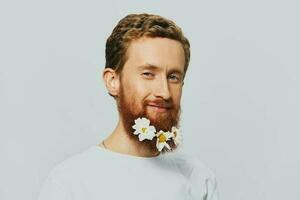 retrato de un gracioso hombre en un blanco camiseta con flores margaritas en su barba en un blanco aislado fondo, Copiar lugar. fiesta concepto y Felicidades. foto