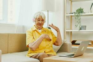 un mayor mujer vistiendo auriculares con un ordenador portátil comunicado en línea por vídeo llamar, sentado en el sofá a hogar y trabajando en un amarillo camisa en frente de un ventana, el estilo de vida de un retirado mujer. foto
