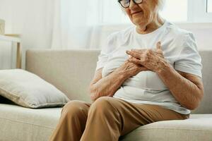 Elderly woman severe chest pain sitting on the sofa, health problems in old age, poor quality of life. Grandmother with gray hair holds her chest with her hands, women's health, breast cancer. photo