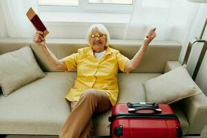 Happy senior woman with passport and travel ticket packed a red suitcase, vacation and health care. Smiling old woman joyfully sitting on the sofa before the trip raised her hands up in joy. photo