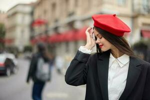 Moda mujer retrato sonrisa dientes en pie en el calle en el ciudad antecedentes en elegante ropa con rojo labios y rojo boina, viajar, cinematográfico color, retro Clásico estilo, urbano Moda estilo de vida. foto