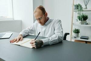 un hombre en un silla de ruedas, un empresario en el oficina trabajos a un computadora portátil, escritura abajo un plan en un computadora portátil, consideración, integración dentro sociedad, el concepto de trabajando un persona con discapacidades foto