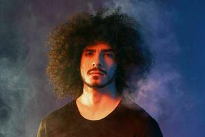 Portrait of a stylish man with curly hair in smoke on a blue background multinational, color light, trend photo