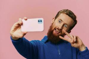 Portrait of a redheaded man with phone in hand taking selfies and photos on his phone with a smile on a pink background, blogger