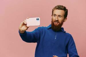 Portrait of a redheaded man with phone in hand taking selfies and photos on his phone with a smile on a pink background, blogger