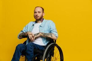 A man in a wheelchair problems with the musculoskeletal system looks at the camera thoughtfully with tattoos on his arms sits on a yellow studio background, health concept a person with disabilities photo