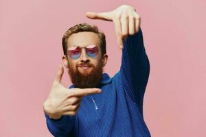 Portrait of a redheaded man wearing sunglasses smiling and dancing, listening to music on a pink background. Hipster with a beard, happiness finger pointing. photo