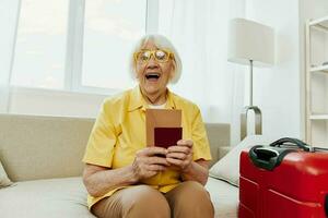 Happy senior woman with passport and travel ticket packed a red suitcase, vacation and health care. Smiling old woman joyfully sitting on the sofa before the trip raised her hands up in joy. photo