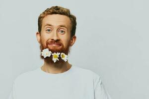 Portrait of a funny man in a white T-shirt with flowers daisies in his beard on a white isolated background, copy place. Holiday concept and congratulations. photo