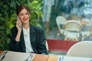 un mujer blogger turista se sienta en un café a un mesa con un teléfono en su manos hablando en el teléfono, móvil comunicaciones y el Internet en un primavera viaje, negocio llamar, Lanza libre trabajo en línea, sonrisa foto